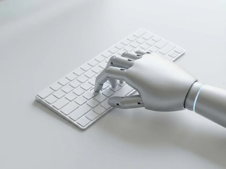 Zilveren robothand drukt op een toets van een wit minimalistisch toetsenbord op een lichtgrijs bureau.