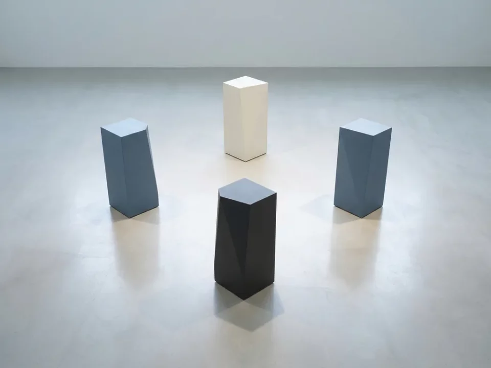 Vier geometrische monolithen in diamantformatie op een gepolijst oppervlak, in leisteenblauw, gebroken wit en antraciet tinten.