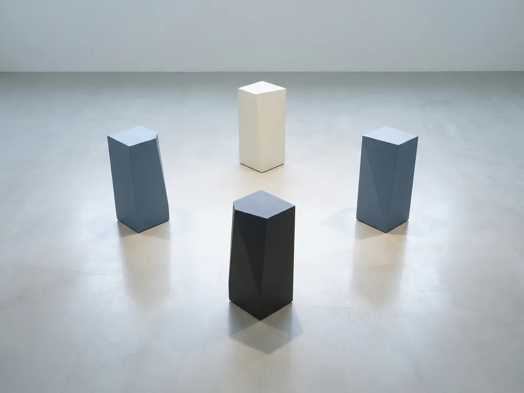 Vier geometrische monolithen in diamantformatie op een gepolijst oppervlak, in leisteenblauw, gebroken wit en antraciet tinten.