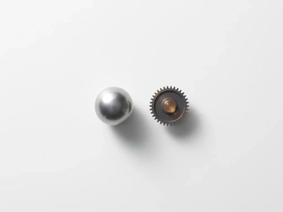 Zilveren metalen bol naast een klein tandwielmechanisme op wit oppervlak, met zachte schaduwen en een vleugje amber.