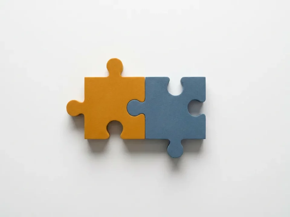 Twee puzzelstukjes in amber en leisteenblauw die in elkaar grijpen op een wit bureau, symbool voor samenwerking en vertrouwen.