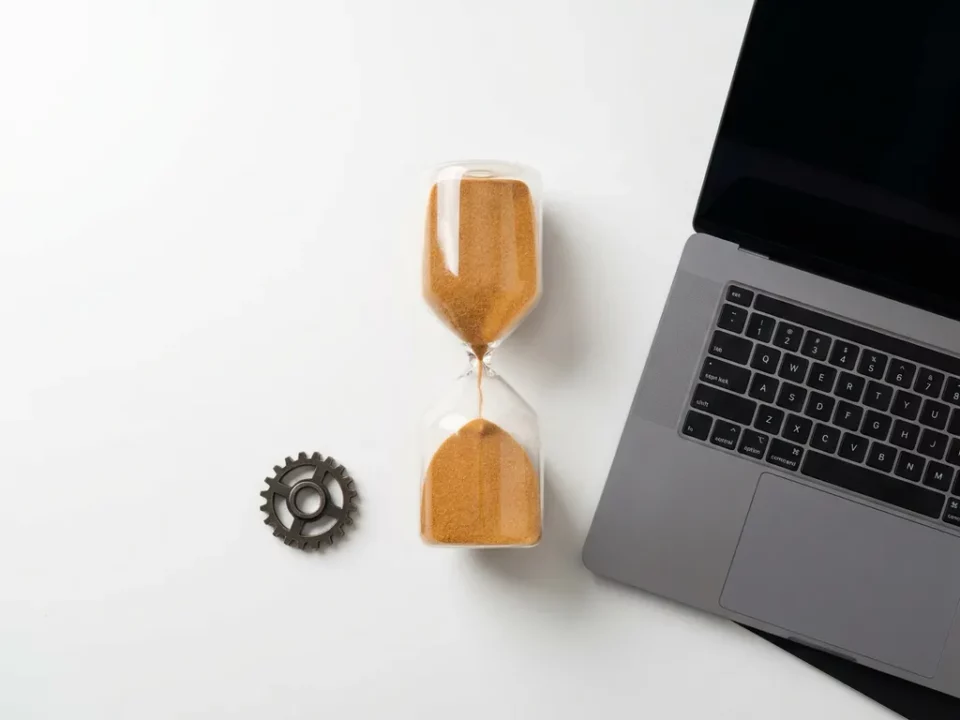 Zandloper met stromend zand naast een open laptop op wit bureau, warme amber tinten, platte bovenaanzicht.